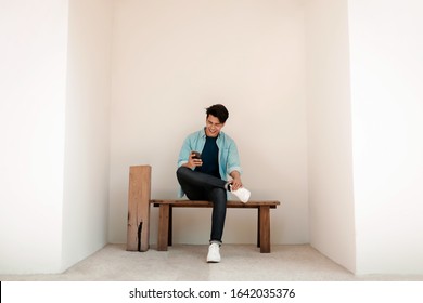 Happy Young Man In Casual Wear Using Mobile Phone While Sitting On Bench By The Wall. Lifestyle Of Modern People. Wide Shot, Full Length