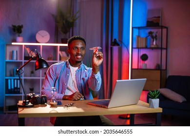 Happy Young Man In Casual Wear Throwing Paper Airplane While Taking Break During Remote Work On Laptop. Smiling Black Guy Sitting At Desk During Evening Time And Having Fun.