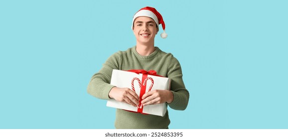 Happy young man in Christmas sweater, with gift box and candy canes on light blue background - Powered by Shutterstock