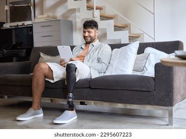 Happy young man with disability leg prosthesis sitting on sofa at home using digital tablet working or elearning, browsing web, searching online. People with amputation disabilities everyday life. - Powered by Shutterstock