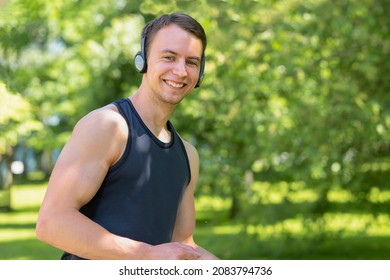 Happy Young Man Doing Workout Outdoor. Muscular Smiling Guy Running In The Park, Training, Listening Music In Headphones. Sport, Fitness Healthy Lifestyle Concept.  