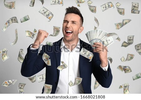 Happy young man with dollars under money rain on light background
