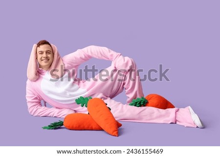 Happy young man in Easter bunny costume lying with plush carrots on lilac background