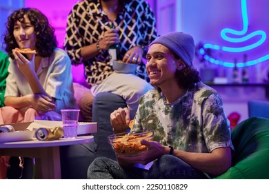 Happy young man eating potato chips and watching movie on tv while sitting against his friends in living room lit by neon light - Powered by Shutterstock