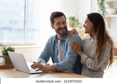 Happy Young Man In Eyeglasses Looking At Laughing Mixed Race Woman, Sitting Together At Wooden Table With Computer. Excited Family Couple Discussing Movie, Having Fun, Communicating Together At Home.