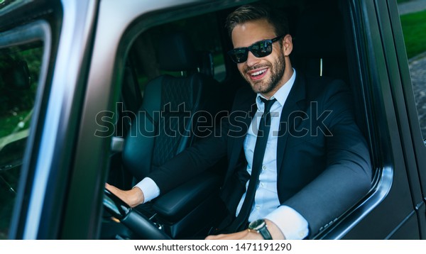 Happy young man in full suit and sunglasses smiling\
while driving a car