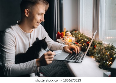 happy young man with funny cat holding credit debt card shopping online purchase xmas presents on laptop home. Christmas gift box, New Year winter sale discount. Safe ordering delivery at quarantine