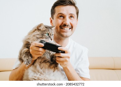 Happy Young Man Gamer Player Having Fun, Holding Gamepad Controller, Playing Video Game While Sitting With Cat At Home On Sofa. Adult Guy With Facial Expression And Pet Enjoying Active Leisure, Hobby.