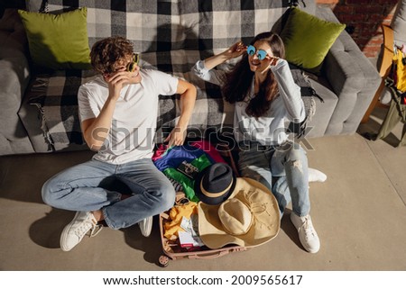 Happy young man and girl, married couple trying on attires, outfits for summer vacation sitting at home, indoors. Look excited, delighted. Concept of relationship, love, lifestyle, lesuire activity.