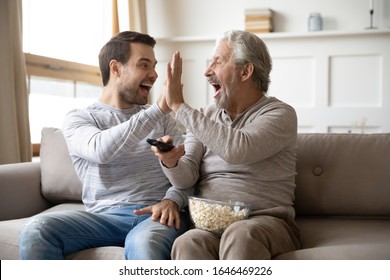 Happy Young Man Giving High Five To Overjoyed Middle Aged Older Father, Celebrating Favorite Team Sport Win At Home. Excited Mature Old Dad Enjoying Watching Championship On Tv With Grownup Son.