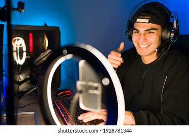 Happy Young Man Giving A Thumbs Up To His Viewers And Friends During A Live Stream With A Smartphone And A Ring Light. Smiling Gamer Playing A Video Game In A Gaming Computer