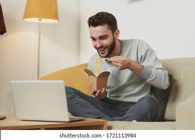 Happy young man in grey sweatshirt and sweatpants eating wok udon with chopsticks from box while watching online movie in front of laptop