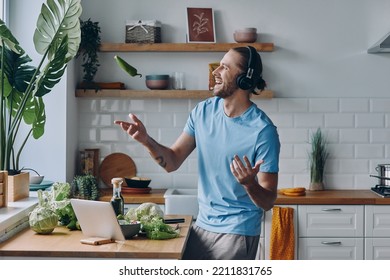 Happy Young Man In Headphones Tossing Up A Green Pepper While Preparing Food At The Kitchen