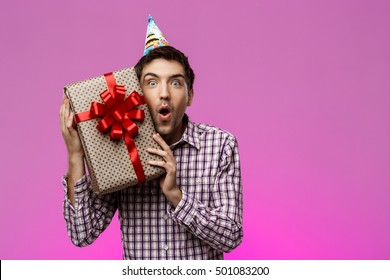 Happy Young Man Holding Birthday Gift In Box Over Purple Background.