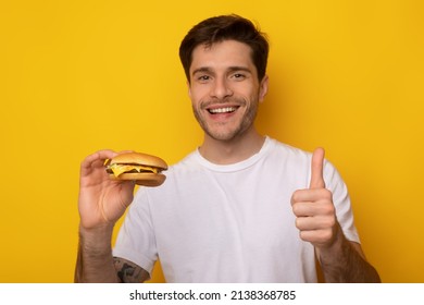 Happy Young Man Holding Burger And Showing Thumbs Up Sign Gesture Approving Hamburger's Taste Smiling To Camera Standing On Yellow Orange Studio Wall, Selective Focus On Hand. I Like Tasty Junk Food