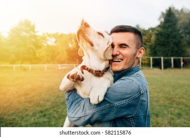 Happy Young Man Holding Dog Labrador In Hands At Sunset Outdoors 