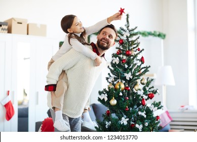 Happy young man holding his daughter on back to help her put star on top of decorated firtree - Powered by Shutterstock