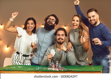 Happy Young Man Holding Pair Of Aces, Surrounded By Group Of Excited, Laughing And Smiling Young Friends, All Looking At Camera, After Successful Game Of Poker At Casino Themed Party At Home Or Club