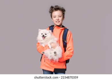 Happy Young Man Holding Pomeranian Dog In Hands On Gray Background In Studio With Copy Space. In An Orange Sweatshirt With A Backpack On His Back, He Smiles Sweetly With His Teeth