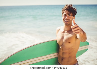 Happy young man holding surfboard showing thumbs up on beach - Powered by Shutterstock