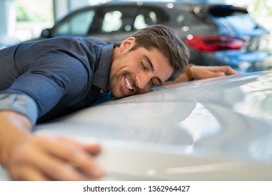 Happy young man hugging his new car in showroom. Satisfied guy with closed eyes embracing the hood of the automobile. Dreaming man lying on car bonnet hugging it.