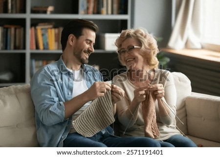 Happy young man learning needlework from smiling older retired mum, sitting together on comfortable couch at home. Joyful two generations family involved in creative handmade activity in living room.