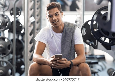 Happy Young Man Listening To Music With Headphones And Looking At Camera. Handsome Guy With Smartphone Sitting On Bench In A Modern Gym. Latin Man Using Phone While Resting After Working Out In Gym.