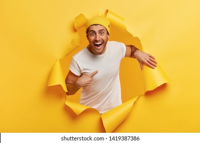 Happy young man points at himself, expresses wonder he was chosen, smiles broadly, wears casual clothes, poses in ripped paper hole, isolated over yellow background, being mentioned or picked