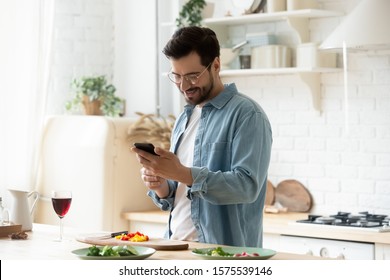 Happy young man preparing romantic dinner searching vegetable recipes diet menu cookbook app using smartphone, smiling husband holding phone cooking healthy vegan food cut salad in kitchen interior - Powered by Shutterstock