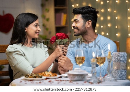 Happy young man proposing girl by giving red roses by arranging candle light dinner - concept of love emotions, wedding present and affectionate