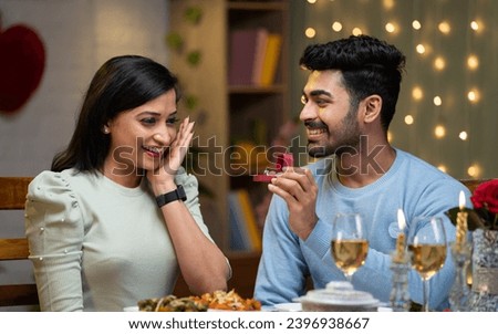 Happy young man proposing to girl with gold ring during candlelight dinner at home - concept of anniversary celebration , engagement and valentines day treat.