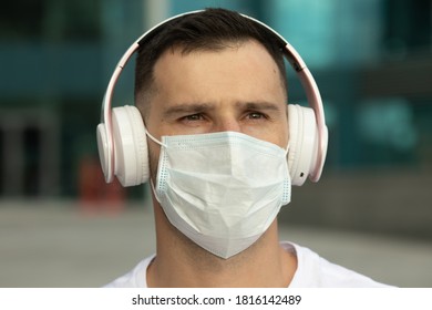 Happy Young Man In Protective Medical Face Mask Listen Music With Wireless Bluetooth Earphones. COVID-19 Coronavirus.