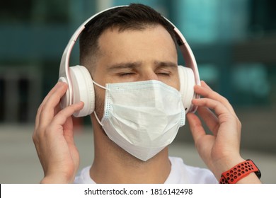 Happy Young Man In Protective Medical Face Mask Listen Music With Wireless Bluetooth Earphones. COVID-19 Coronavirus.