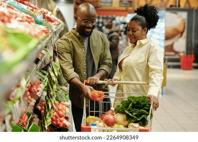 Happy young man putting fresh pepper into shopping cart in supermarket while standing between display with vegetables and his wife - Powered by Shutterstock