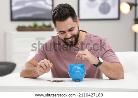 Happy young man putting money into piggy bank at white table indoors