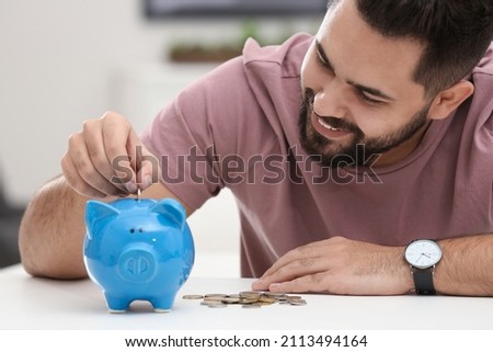 Happy young man putting money into piggy bank at white table indoors