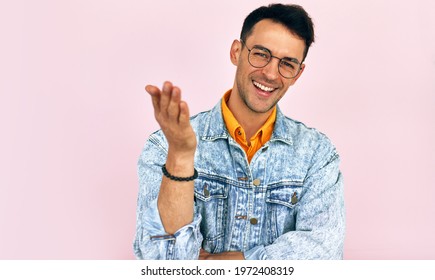 Happy Young Man Raises His Hand During A Positive Conversation With Someone. Male In A Denim Jacket And Transparent Spectacles Pulls Arm In Welcome Gesture, Isolated Over Pink Background.