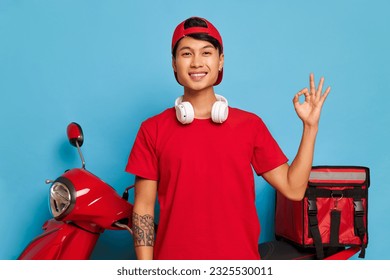 Happy young man in red cap and red t-shirt with earphones on his neck posing near red scooter on blue background, shows ok sign with one hand, nice drive concept, copy space - Powered by Shutterstock