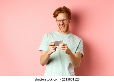 Happy Young Man With Red Messy Haircut, Wearing Glasses, Playing Video Game On Smartphone And Having Fun, Looking At Mobile Screen, Standing Over Pink Background