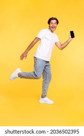 Happy Young Man Showing Empty Smartphone Screen While Jumping Up Over Yellow Studio Background, Full Body Length Shot. Excited Running Guy Recommending Nice And Useful Mobile Application