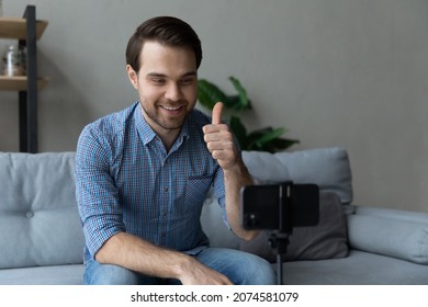 Happy young man showing thumbs up gesture recording video on cellphone web camera standing on stabilizer on table, recommending service enjoying blogging activity at home, streaming stories online.