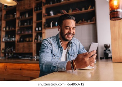 Happy Young Man Sitting At Coffee Shot With Earphones Making Video Call To Friend. Caucasian Male Having Video Chat On Smart Phone At Cafe.