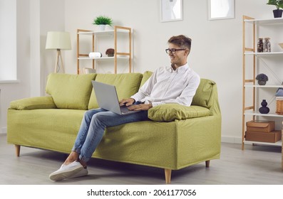 Happy young man sitting with his notebook PC on sofa at home. Remote student or corporate employee working on modern laptop computer while resting on comfortable couch in living room - Powered by Shutterstock