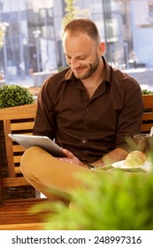 Happy Young Man Sitting On Bench, Using Tablet In Outdoor Cafe.