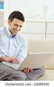 Happy Young Man Sitting On Couch And Working On Laptop Computer At Home, Smiling.