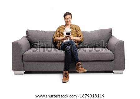 Happy young man sitting on a gray sofa and typing on a mobile phone isolated on white background