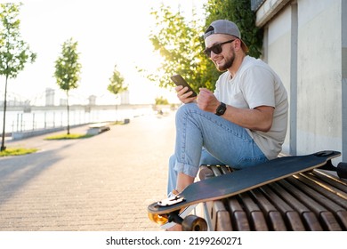 Happy Young  Man With Skateboard Looking At Mobile Phone Social Media App To Connect With Friends. Urban Lifestyle, Street Fashion And Technology Concept.