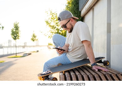 Happy Young  Man With Skateboard Looking At Mobile Phone Social Media App To Connect With Friends. Urban Lifestyle, Street Fashion And Technology Concept.