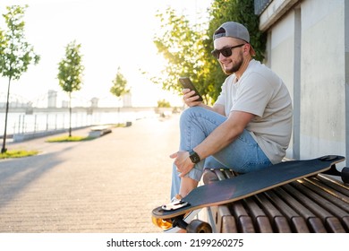 Happy Young  Man With Skateboard Looking At Mobile Phone Social Media App To Connect With Friends. Urban Lifestyle, Street Fashion And Technology Concept.