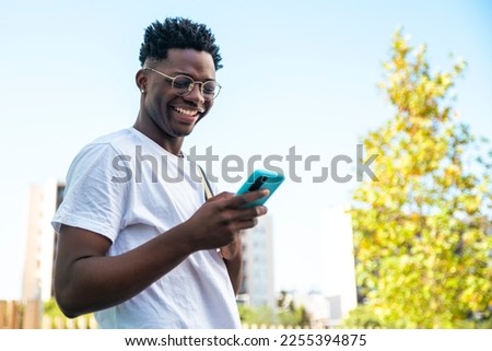 Happy young man smiling and looking his smartphone with a backpack in the park - Low angle portrait of a handsome young man - education concept